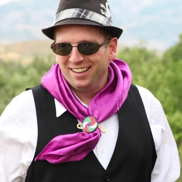 Smiling man wearing a white long-sleeve shirt, black vest, and black fedora with a purple silk scarf around his neck.